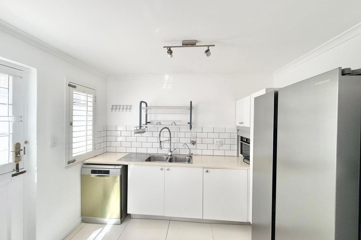 A clean, functional kitchen featuring white cabinetry, subway tile backsplash, stainless steel sink, and built-in oven with tiled flooring and plantation shutters.
