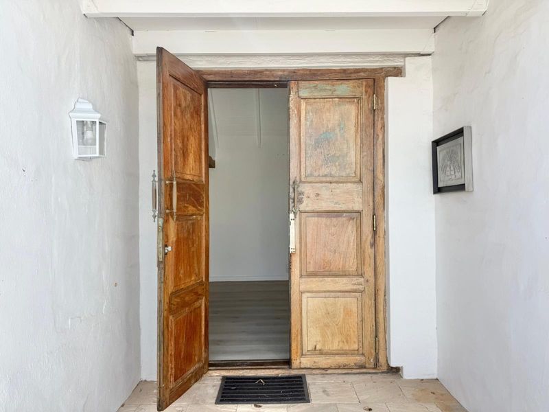 A rustic double wooden entrance door leading into a bright interior space, featuring a classic exterior wall lantern and tiled porch flooring.