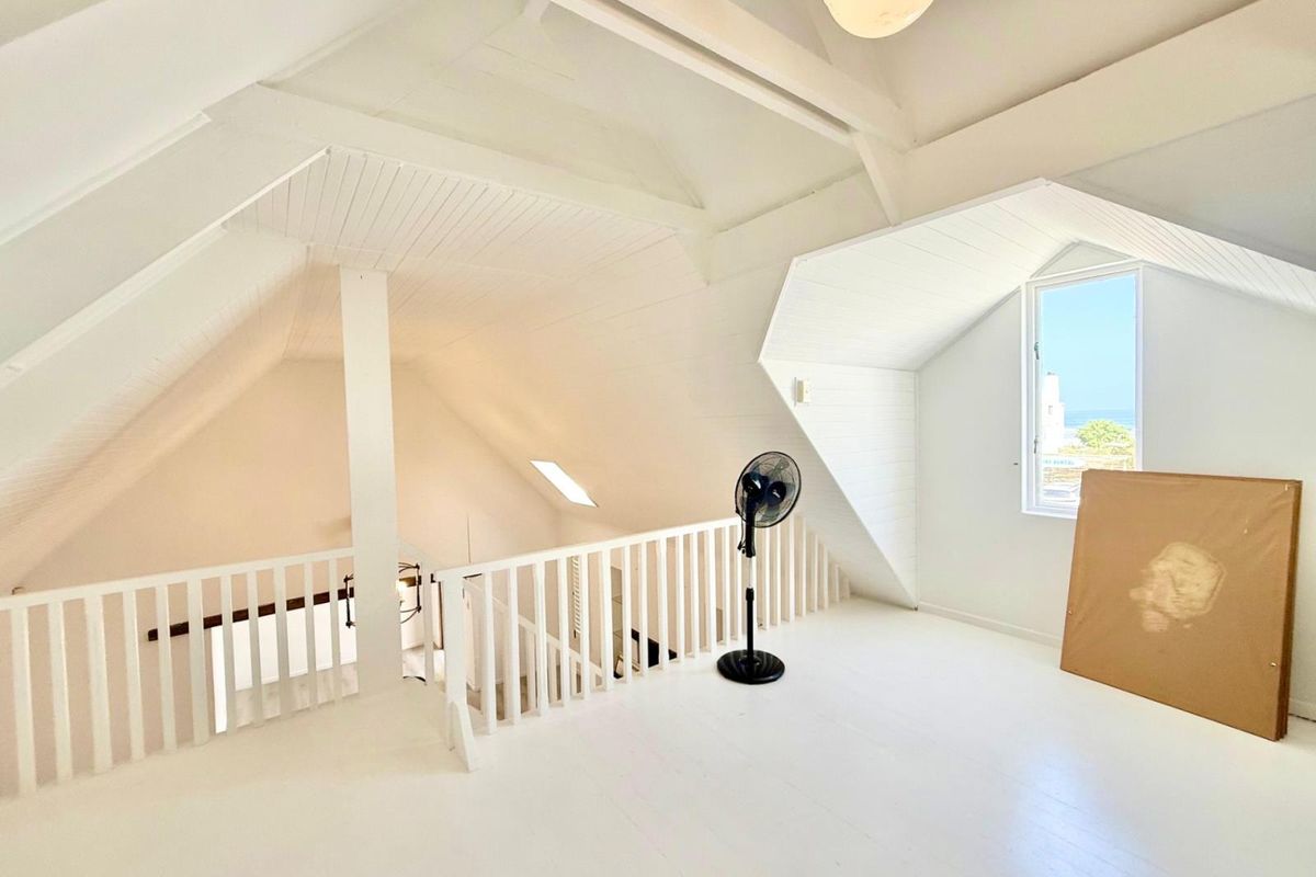 Spacious attic-style bedroom featuring vaulted ceilings, exposed timber beams, a protective balustrade, and a dormer window with coastal views.