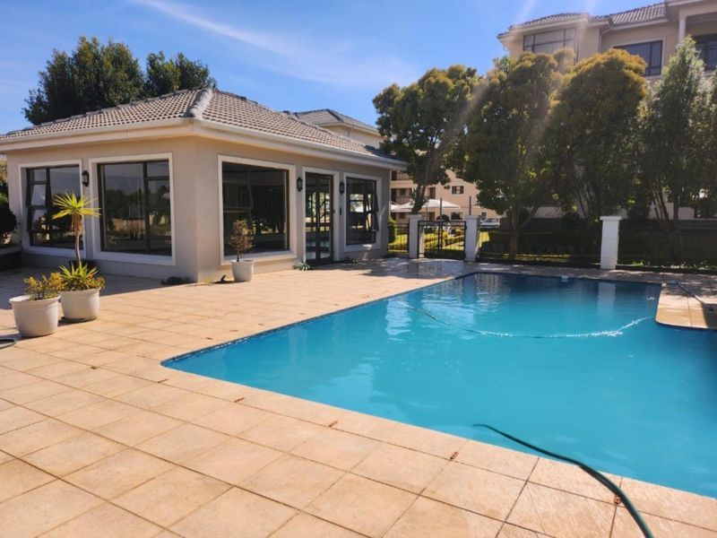 A well-maintained outdoor pool area featuring a tiled deck and a detached structure with large windows, set within a secure residential complex.