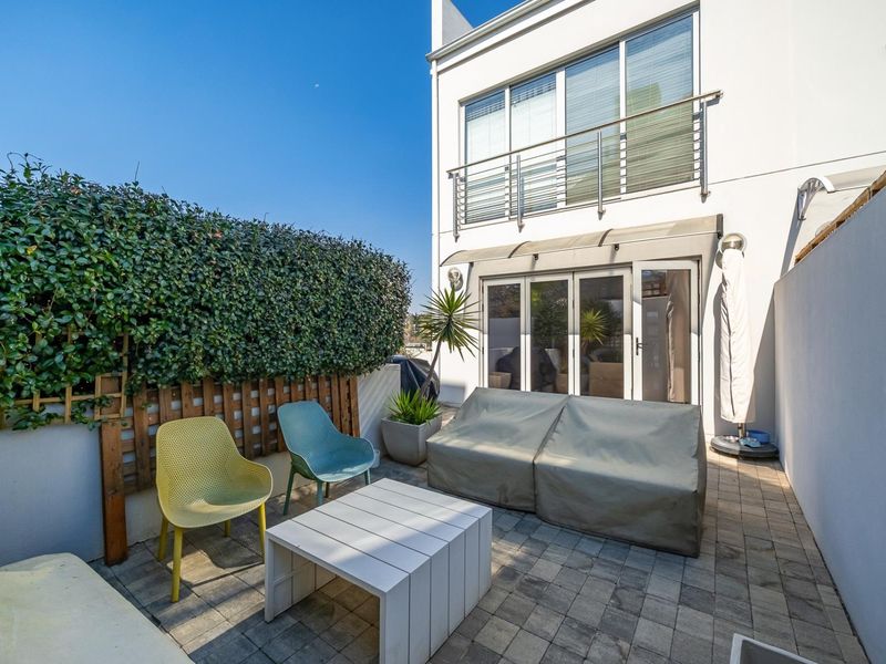 A private paved patio area featuring bi-fold doors leading into the townhouse, complemented by a balcony above and established hedge screening.