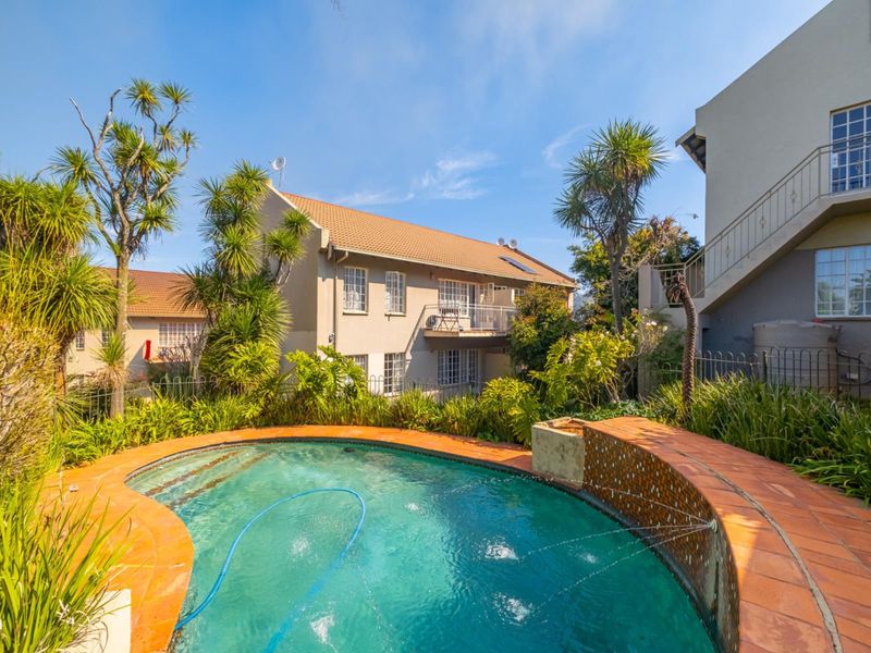 A well-maintained outdoor pool area featuring a tiled surround, water feature, and landscaped garden, set against the rear elevation of the townhouse complex.
