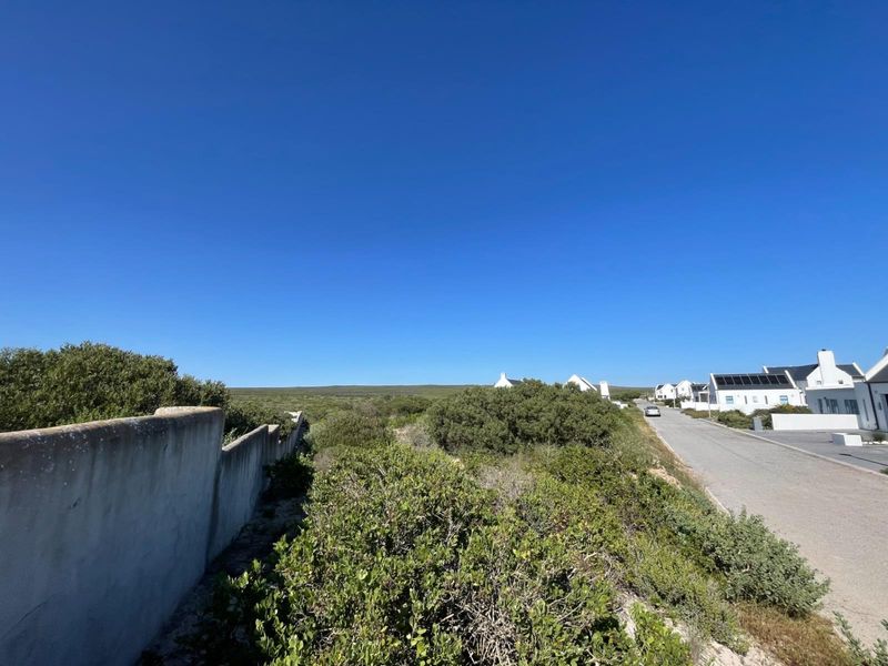 A street view showing the neighbourhood approach, asphalt road, and surrounding coastal landscape with nearby residential properties featuring solar panels.