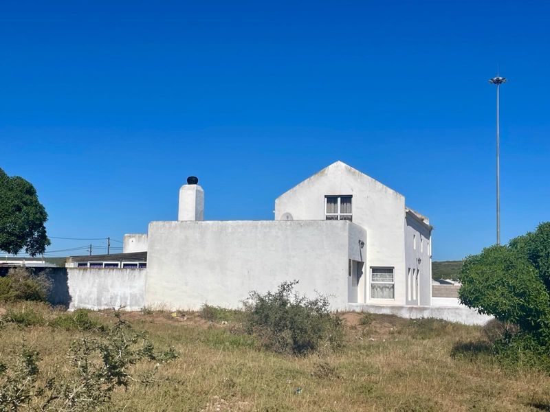 Rear view of a white-painted house featuring a prominent chimney, gabled roof, and surrounding boundary wall set against a clear blue sky.