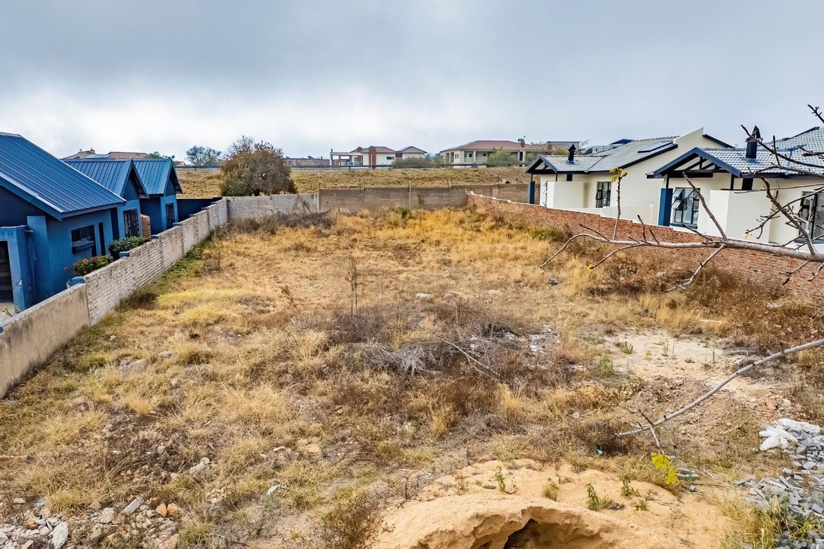 A vacant residential plot situated between existing housing developments, featuring boundary walls and natural ground cover.