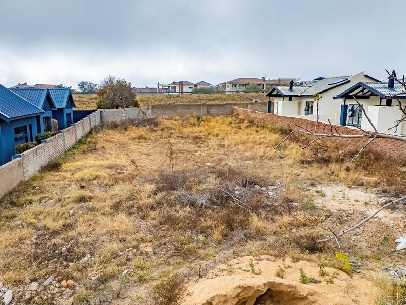 A vacant residential plot situated between existing housing developments, featuring boundary walls and natural ground cover.