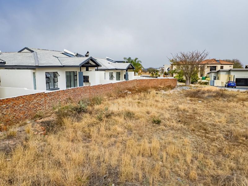 A view of a vacant residential plot adjacent to established housing, featuring a brick boundary wall and surrounding residential infrastructure.