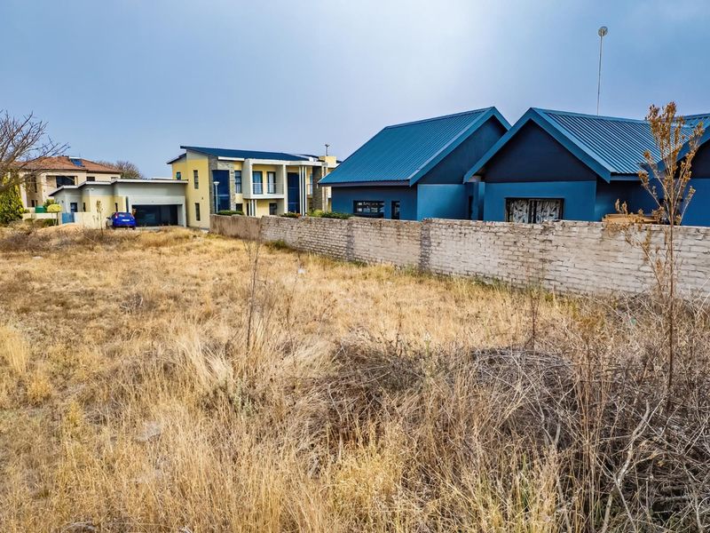A view of a vacant residential plot featuring dry, overgrown grass and a brick boundary wall, situated in a developed neighborhood.