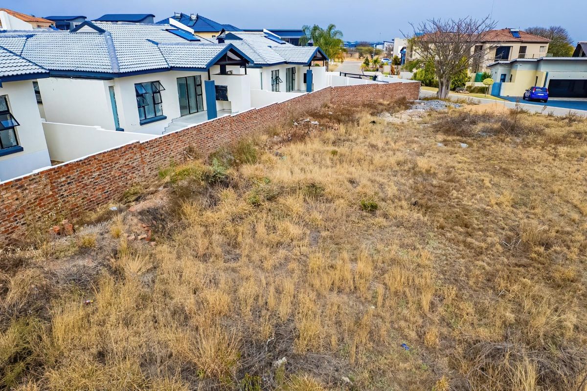 Aerial view of a vacant residential plot adjacent to an established housing development, featuring a brick boundary wall and natural ground cover.