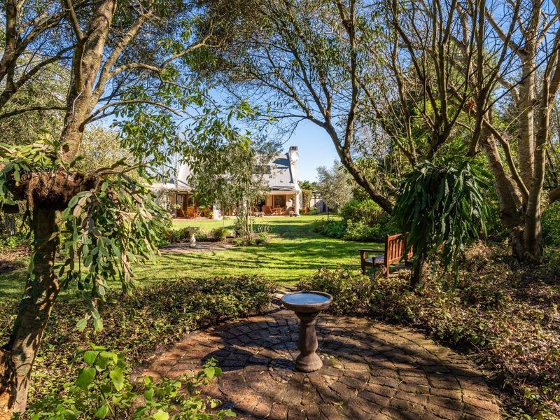 A lush, well-maintained rear garden featuring mature trees, a manicured lawn, and a circular brick-paved patio area leading toward the house.