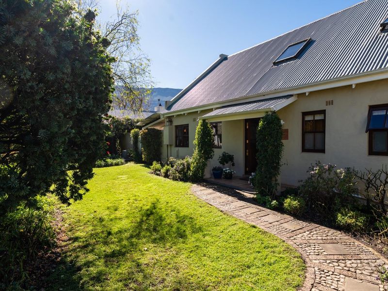 A well-maintained rear exterior view featuring a corrugated metal roof with skylights, a charming cobblestone pathway, and a lush, landscaped garden.