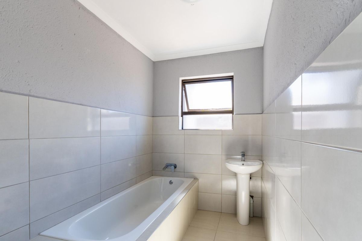 A clean, minimalist bathroom featuring a white bathtub, pedestal basin, and neutral wall and floor tiling with a functional top-hung window.