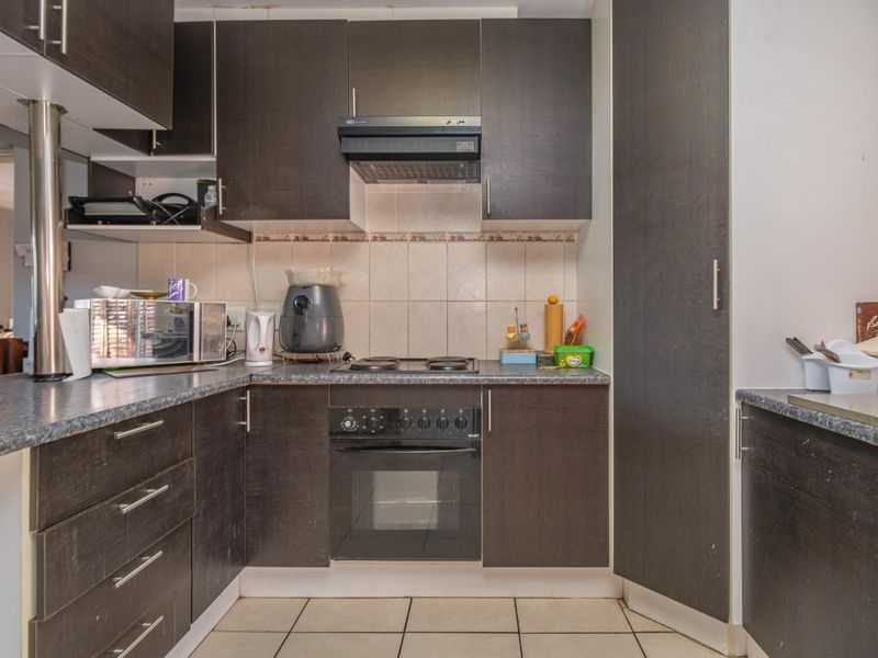 A compact kitchen featuring dark laminate cabinetry, built-in oven with electric hob, extractor fan, tiled backsplash, and tiled flooring.
