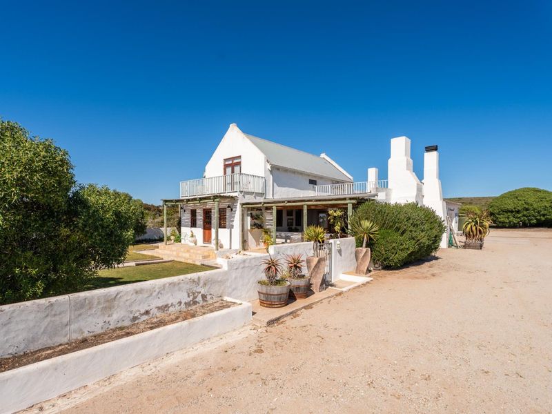 A charming white farmhouse with a metal roof, featuring a covered patio, upper-level balcony, and multiple chimneys, set on a spacious rural property.