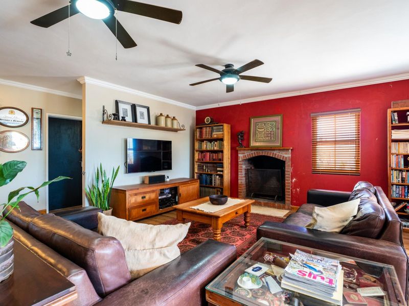 A cozy living room featuring a brick fireplace, dual ceiling fans, and wooden window blinds, offering a warm and traditional atmosphere.