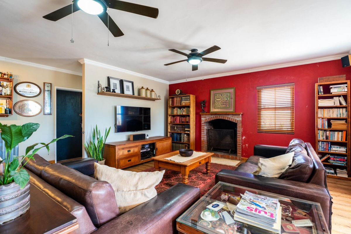 A cozy living room featuring a brick fireplace, dual ceiling fans, and wooden window blinds, offering a warm and traditional atmosphere.