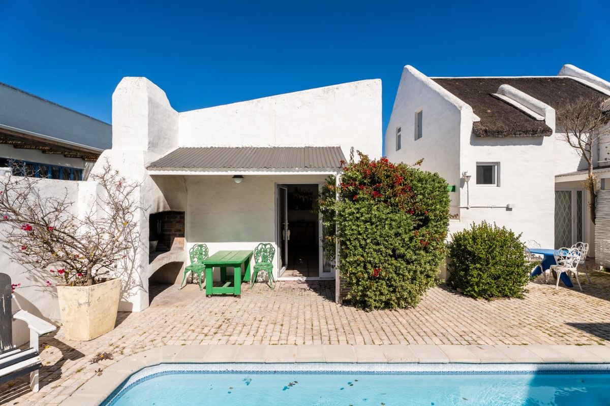 Rear exterior view featuring a paved patio area with a built-in braai, adjacent to a swimming pool and white-walled residential structures.