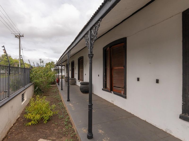 A covered veranda featuring classic wrought iron columns and timber shutters, overlooking a side garden area with perimeter security fencing.