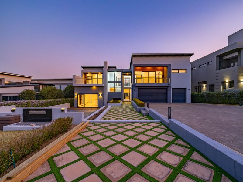 Modern double-story residence featuring a paved driveway, double garage, and upper-level balconies with glass balustrades, set within a well-maintained landscaped estate.