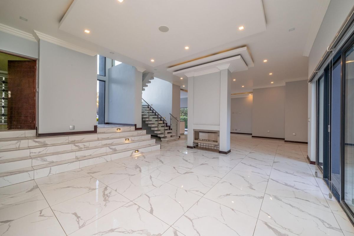 Spacious open-plan living area featuring polished marble-look floor tiles, a central built-in fireplace, recessed ceiling lighting, and a modern staircase with glass balustrades.