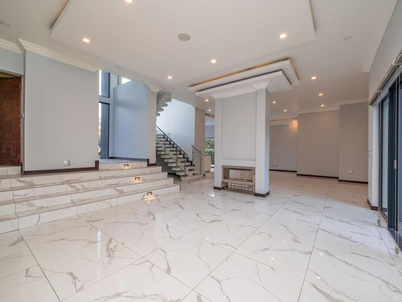 Spacious open-plan living area featuring polished marble-look floor tiles, a central built-in fireplace, recessed ceiling lighting, and a modern staircase with glass balustrades.