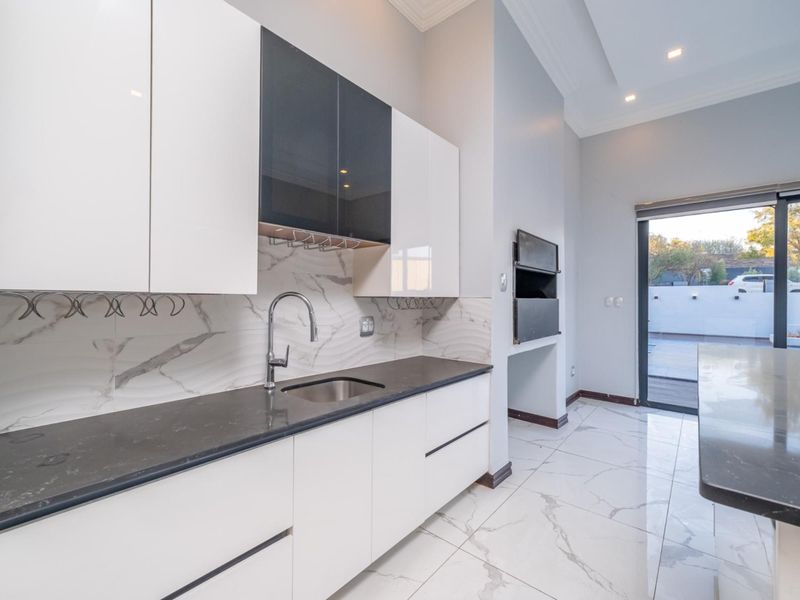 Modern kitchen featuring high-gloss cabinetry, stone countertops, marble-look tile backsplash, and a built-in braai, with direct access to the outdoor patio area.