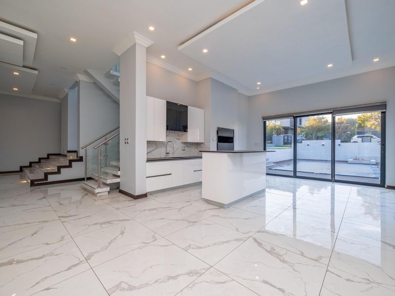 Modern open-plan living and kitchen area featuring high-gloss cabinetry, marble-look floor tiles, and a sleek staircase with glass balustrades leading to the upper level.