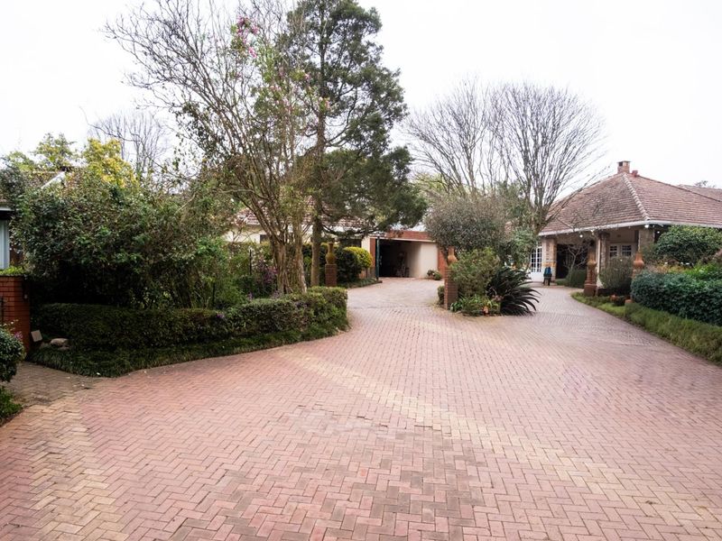 A spacious front exterior view featuring a large paved driveway, mature landscaped gardens, and a traditional brick house with a tiled roof.