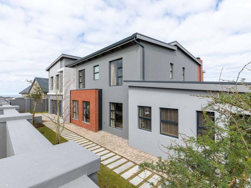 A modern double-storey residence featuring a combination of grey plastered walls and red brick accents, complemented by a paved walkway and boundary wall.