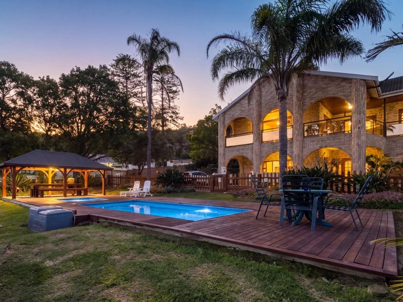 A beautifully landscaped pool area featuring a swimming pool, timber decking, and a covered gazebo, set against a multi-story brick guest house.