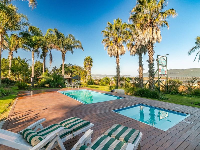 A well-maintained outdoor pool area featuring dual swimming pools set within timber decking, surrounded by mature palm trees and landscaped garden space.
