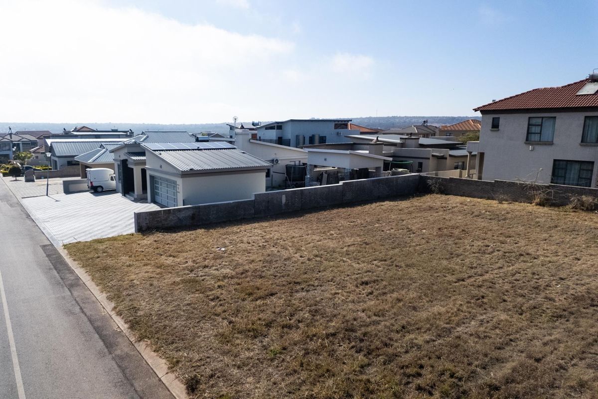 Aerial view of a vacant residential plot situated in a developed neighborhood, featuring a boundary wall and adjacent paved street access.