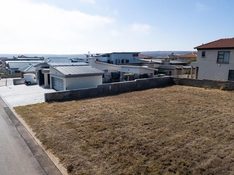 Aerial view of a vacant residential plot situated in a developed neighborhood, featuring a boundary wall and adjacent paved street access.