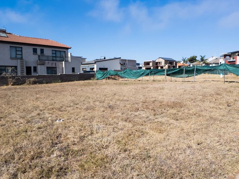 A vacant residential plot of land featuring dry grass, a perimeter boundary wall, and temporary green shade cloth fencing.