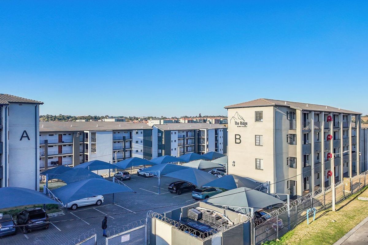 Aerial view of The Ridge apartment complex featuring multi-story residential buildings, paved parking areas with shade sails, and perimeter security fencing.