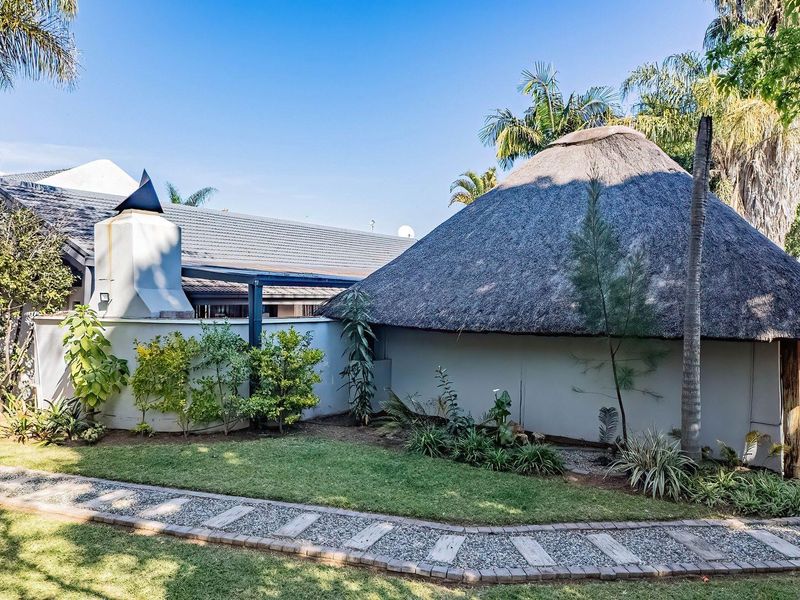 A well-maintained rear garden area featuring a unique thatched-roof structure, an outdoor fireplace, and a paved walkway set within a landscaped yard.