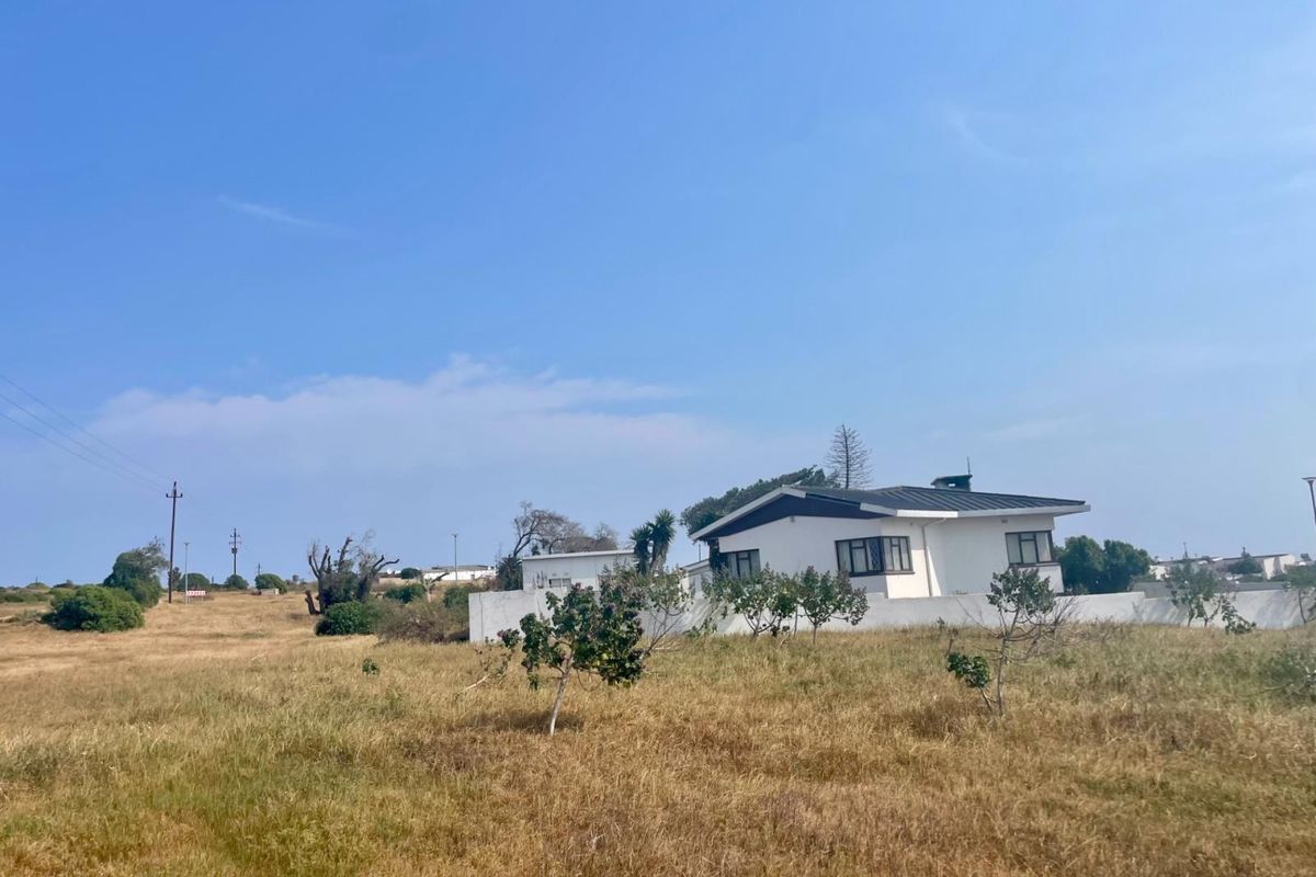 A view of a residential property featuring a white exterior facade, boundary wall, and pitched roof set within a large, open grassed plot.
