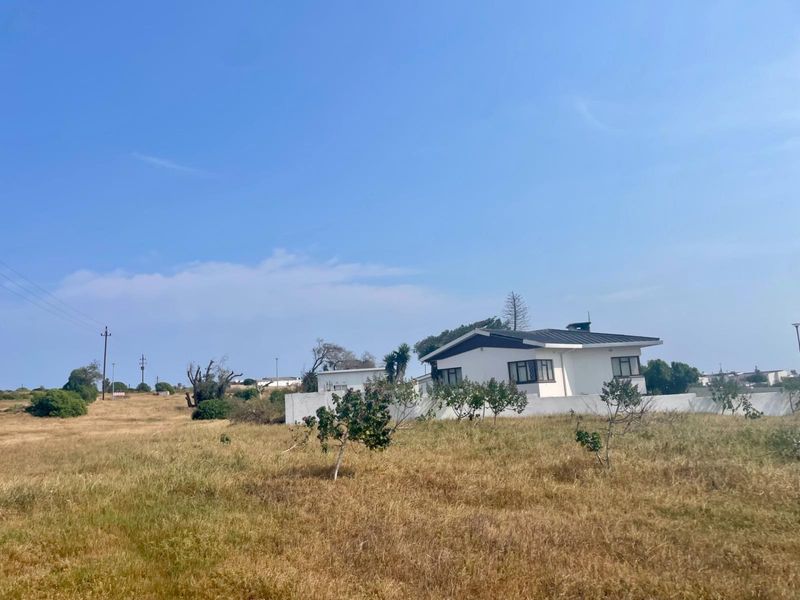 A view of a residential property featuring a white exterior facade, boundary wall, and pitched roof set within a large, open grassed plot.