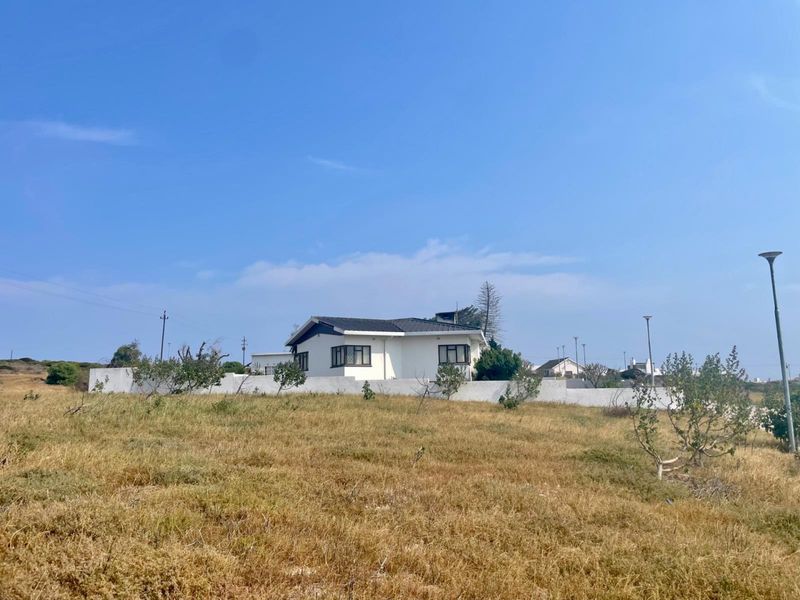 A view of a residential property featuring a white exterior, pitched roof, and surrounding boundary wall, set within a large open plot.