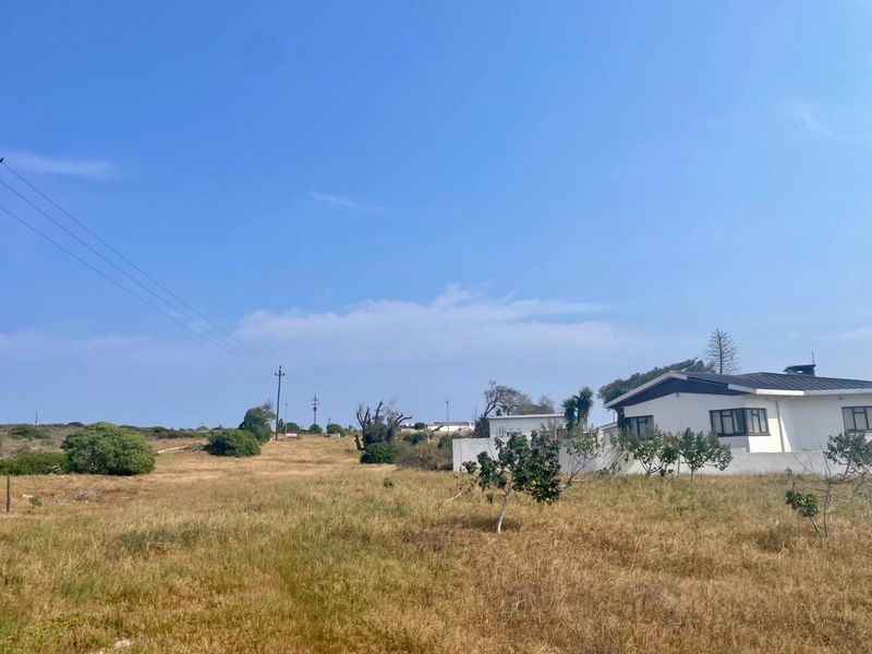 A view of a residential vacant land plot featuring dry grass, power lines, and a partial view of an adjacent white building.