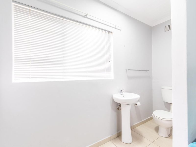 A clean, functional bathroom featuring a pedestal basin, toilet, tiled flooring, and natural light provided by a window with venetian blinds.