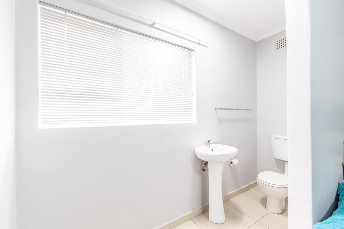 A clean, functional bathroom featuring a pedestal basin, toilet, tiled flooring, and natural light provided by a window with venetian blinds.