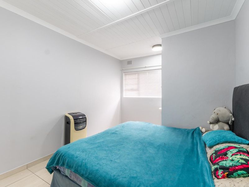 A functional bedroom featuring tiled flooring, a tongue and groove ceiling, and a window fitted with venetian blinds.