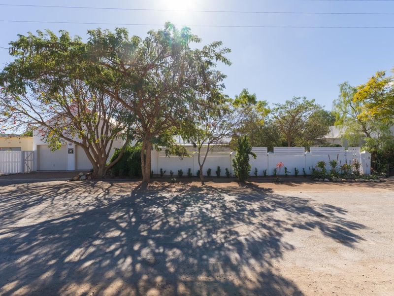 Front view of the property featuring a white boundary wall, gated entrance, and garage, set behind mature trees.