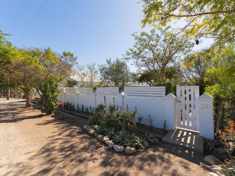 A well-maintained front boundary wall with a white pedestrian gate, landscaped garden beds, and mature trees along a quiet street.