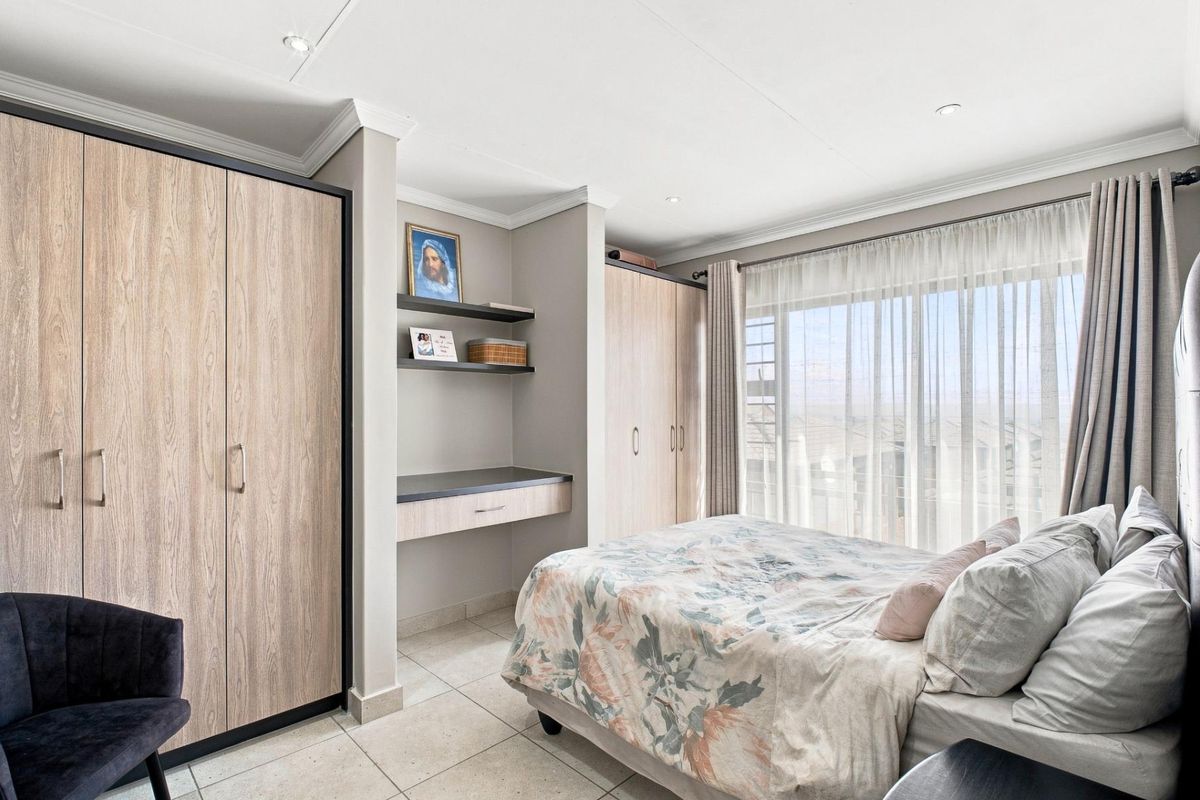 A well-maintained bedroom featuring built-in wooden cupboards, a integrated desk area, tiled flooring, and large windows with curtain rails.