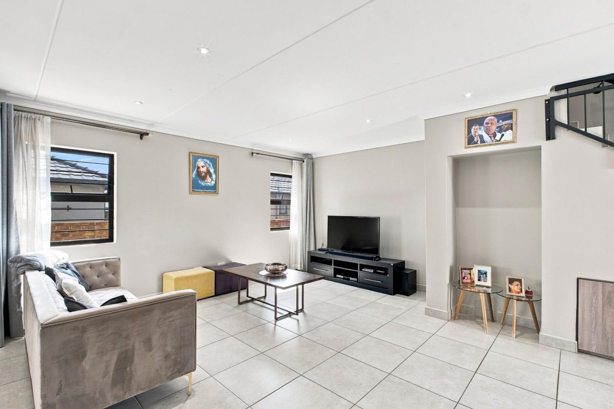 A bright and spacious living room featuring neutral tiled flooring, recessed downlighting, and a built-in staircase alcove.