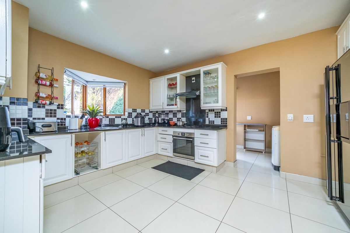 A well-lit kitchen featuring white cabinetry, granite countertops, a tiled backsplash, and a bay window, with an adjacent scullery area.
