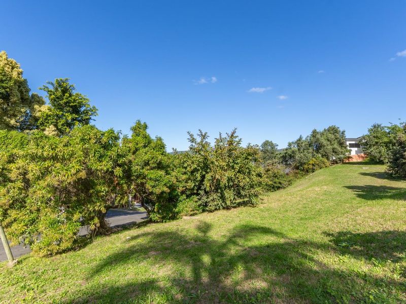 A view of a sloped, grassed residential plot featuring mature trees and established landscaping, offering a natural setting for potential development.