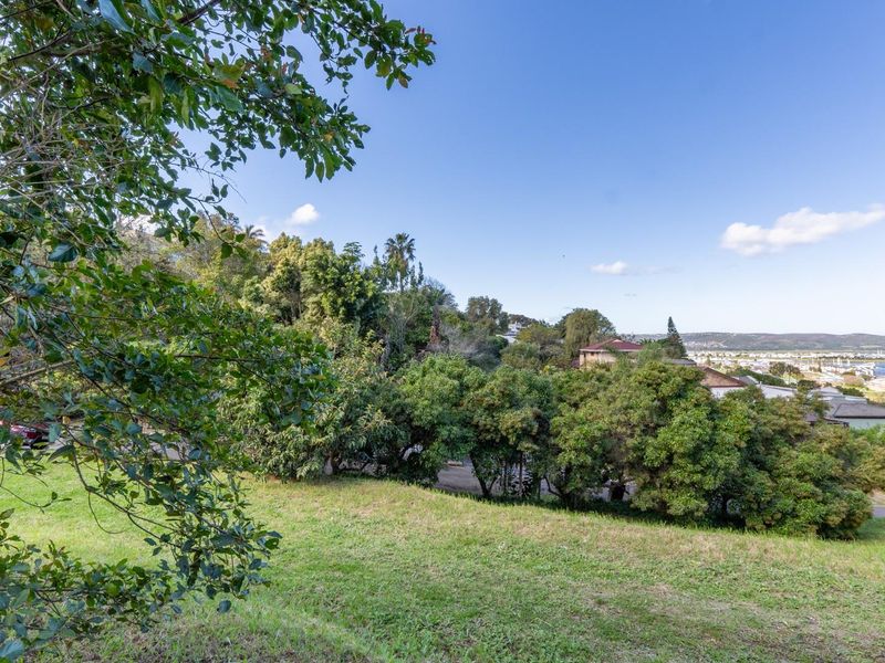 A scenic view overlooking a sloped residential plot with mature trees and distant coastal landscape.